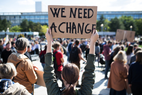 Eine Demonstration, eine Frau hält in einer Menschenmenge ein Plakat hoch auf dem steht "We need a change"