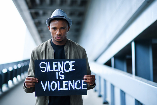 Ein dunkelhäutiger junger Mann, mit Hut, hält ein Plakat in den Händen, auf dem steht "Silence is violence"