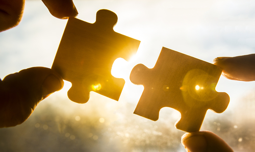 Zwei Puzzleteile werden passend von je zwei Fingern zusammengehalten, im Hintergrund erkennt man einen Sonnenuntergang.