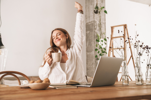 Frau sitzt mit Kaffee in einer Hand in Siegerpose vor dem Laptop.