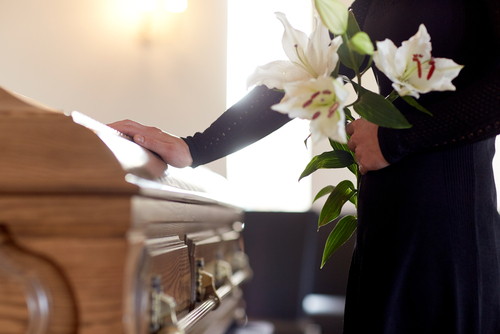 Beerdigung, Abschied nehmen. Frau steht mit Lilien im Arm neben Sarg.