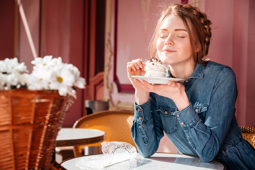 Rothaarige Frau sitzt am Tisch von einem Café, hält Teller in der Hand und riecht an Muffin