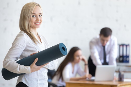 Business Frau mit Trainings-Matte unterm Arm. Im Hintergrund sind 2 Kollegen am Arbeiten.