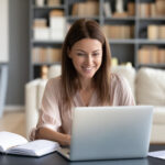 Braunhaarige Frau sitzt am Schreibtisch vor Laptop, neben ihr liegt ein Notizbuch