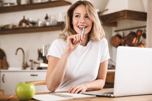 Blonde Frau sitzt vor Küche am Laptop und überlegt. Sie hält einen Stift zum Mund.