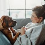 Frau sitzt mit braunem Hund auf Sofa. Tierkommunikation