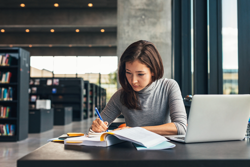 Frau sitzt in der Bibliothek am Tisch, sie schreibt. Neben ihr liegen Bücher und Notebook