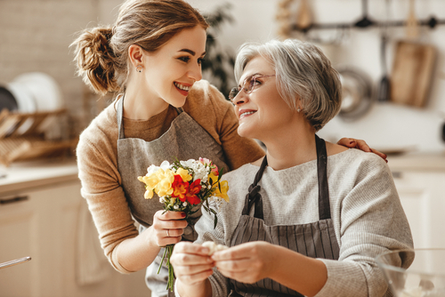 Tochter steht neben sitzender Mutter, und schenkt ihr Blumen. Beide lächeln