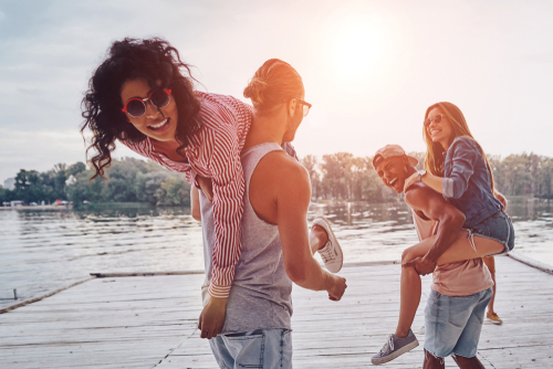 4 Freunde am See, die zusammen Spaß haben. Die Männer tragen die Frauen und lachen