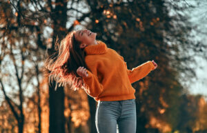 Dunkelhaarige Frau mit orangen Pulli tanzt im Herbst im Freien. Sie lächelt und blickt nach oben.