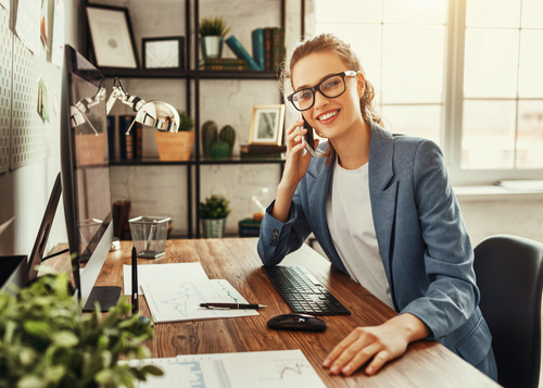 Blonde Frau mit Brille sitzt vor PC am Schreibtisch und telefoniert