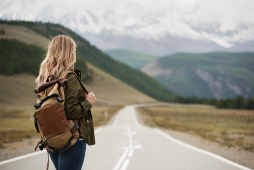Blonde haarige Frau mit Rucksack steht auf Landstraße, man sieht nur ihren Rücken.