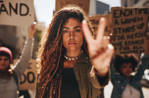 Frau mit lockigem Haar zeigt bei einer Demonstration für Frauenrechte das Peace-Zeichen.