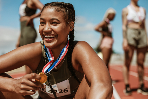 Sportlerin mit Zöpfen sitzt lachend auf dem Boden der Laufbahn im Stadion und hält eine Goldmedaille in der Hand.